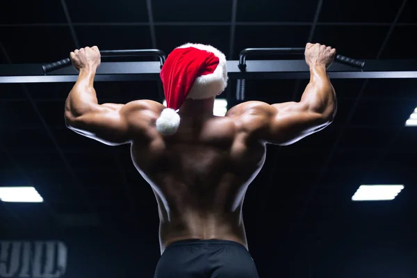 Giovane sexy Babbo Natale in un cappello di Natale sta lavorando fuori pompaggio muscoli della schiena in palestra guadagnando peso sulle macchine — Foto Stock