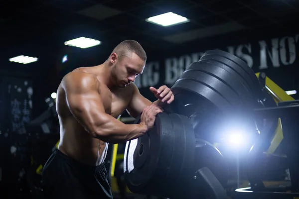 Joven atleta muscular hombre de apariencia modelo está descansando después de ejercitar los músculos del entrenamiento en el gimnasio de pie junto a los discos —  Fotos de Stock