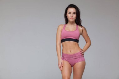 Pembe spor kıyafetli çekici genç esmer kadın gri arka planda yalnız poz veriyor.