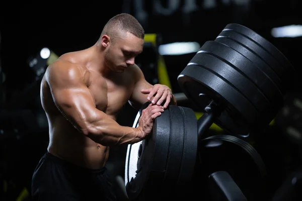 Joven atleta muscular hombre de apariencia modelo está descansando después de ejercitar los músculos del entrenamiento en el gimnasio de pie junto a los discos —  Fotos de Stock