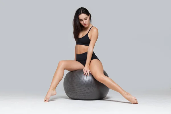Full length portret van sportieve sexy meisje zitten op fitness bal stretching haar been uit geïsoleerd op grijze achtergrond — Stockfoto
