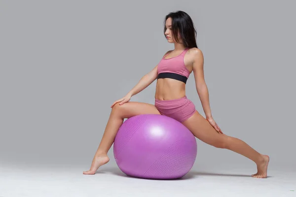 Menina sexy no topo rosa e shorts executa um exercício de alongamento usando um fitball isolado no fundo cinza — Fotografia de Stock