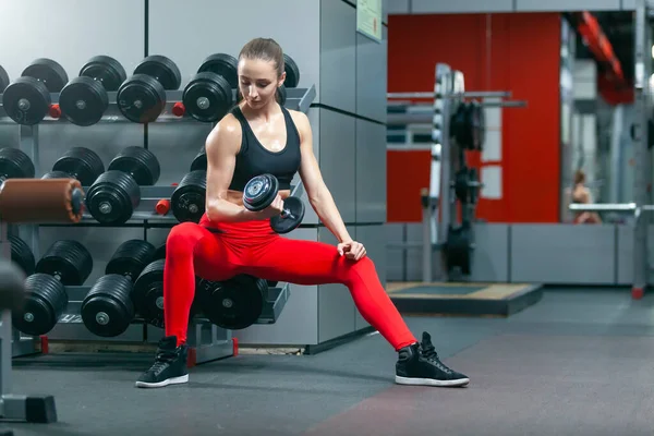 Fiatal izmos nő csinál koncentrált emelés bicepsz egyik kezét súlyzóval, miközben ül a padon az edzőteremben — Stock Fotó