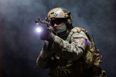 Askeri kamuflaj üniformalı, miğferli, gözlüklü ve maskeli asker portresi tüfeği yüzüne yakın tutuyor ve saldırı pozisyonunda, siyah arka planda duman altında durarak nişan alıyor.