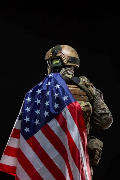 Visão traseira de um militar com um chapéu duro e equipamento completo, coberto sobre o ombro da bandeira da América isolado em preto. O conceito de patriotismo, a proteção da Pátria, o amor pelo país — Fotografia de Stock