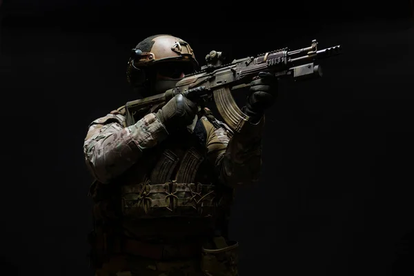 Vista lateral de um militar vestido com equipamento e um capacete, que aponta uma metralhadora com visão de colimador, pronta para atirar para matar, isolada em um fundo preto — Fotografia de Stock