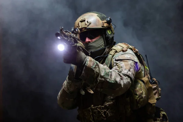 Ritratto di soldato dell'esercito in uniforme militare mimetica, casco, occhiali e maschera tiene il fucile vicino al volto e prende di mira usando la vista del collimatore in piedi in posizione di attacco, in fumo su sfondo nero — Foto Stock