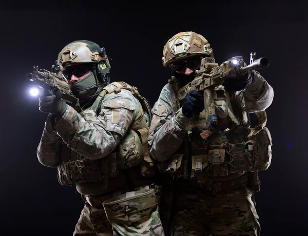Two soldiers in military gear and bulletproof vests cover each other and raise their submachine guns to their faces taking aim, in full combat readiness, isolated on black background — Stock Photo, Image