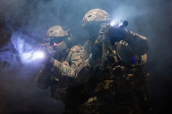 Két katona katonai felszerelésben és golyóálló mellényben befedik egymást, és felemelik géppisztolyaikat, célba véve őket, teljes harckészültségben áttörik a füstöt. — Stock Fotó