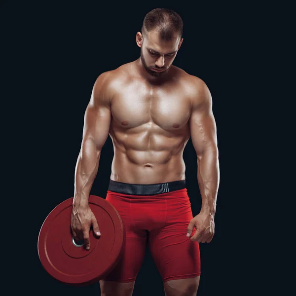 Foto de atlético joven con el torso desnudo y buen físico con disco de peso pesado sobre fondo negro —  Fotos de Stock