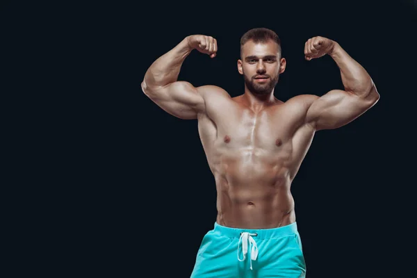 Front view of muscular and fit young bodybuilder fitness male model demonstrating his biceps isolated on black background — Stock Photo, Image