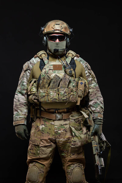 Солдат спецназа в военной экипировке, защитной маске, очках и маске прячет лицо с автоматом стоит прямо вверх, глядя на камеру, изолированную на черном фоне в студии