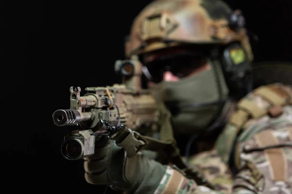 Detailní záběr vojáka v maskáčích a plné vojenské výstroji, v helmě, brýlích a ochranné masce, zvedne pušku k obličeji a míří, připravuje se k výstřelu, izolovaný na černém pozadí — Stock fotografie