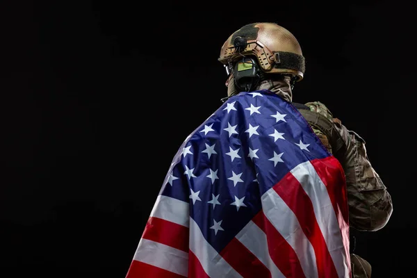 Vista trasera de un militar con un sombrero duro y equipo completo, cubierto sobre el hombro de la bandera de Estados Unidos aislado en negro. El concepto de patriotismo, la protección de la Patria, el amor por el país — Foto de Stock