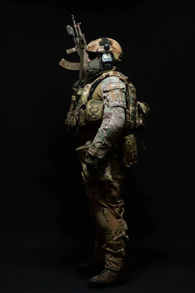 Visão lateral de comprimento total retrato de um soldado do exército de pé em pleno uniforme militar, vestindo um colete à prova de balas, capacete, óculos e máscara, segurando uma submetralhadora isolada em um fundo preto — Fotografia de Stock