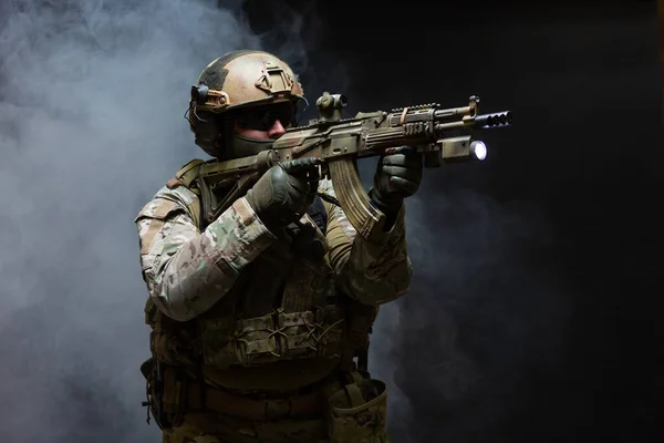 Vista lateral do soldado do exército em uniforme de camuflagem militar, capacete, óculos e máscara segura rifle perto do rosto e mira usando visão de colimador em posição de ataque, em fumaça no fundo preto — Fotografia de Stock