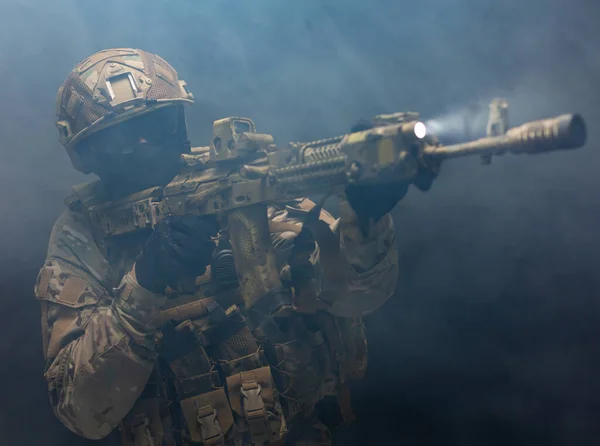 Foto vista frontal do soldado homem em verde desgaste militar vestindo colete de descarga, balaclava, óculos, capacete segurando arma automática tendo objetivo não tem medo de perigo, de pé sozinho em fumaça — Fotografia de Stock