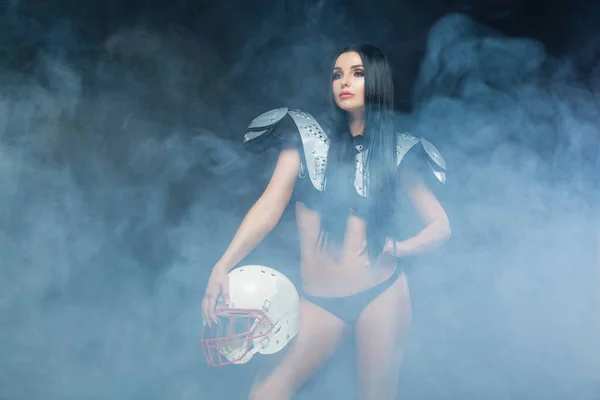 American Football. Vorderseite der jungen sportlichen Brünette trägt sexy Uniform eines Rugby-Fußballers posiert mit Helm in Rauch isoliert auf schwarzem Hintergrund — Stockfoto