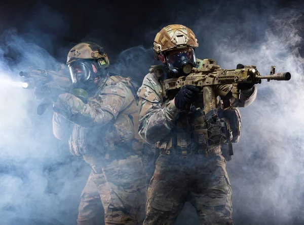 Twee militairen in militaire uitrusting, kogelvrije vesten en gasmaskers bedekken elkaar en richten hun geweren op elkaar, in volledige gevechtsbereidheid om door de rook van chemische wapens te breken. — Stockfoto