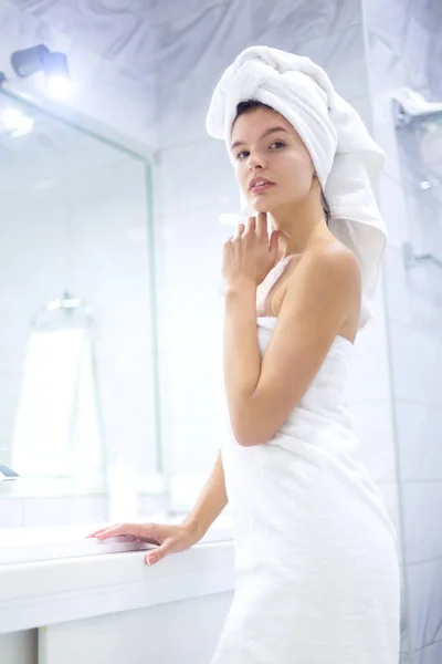 Retrato de una joven feliz y sonriente mujer de pie en el baño con toallas blancas envueltas alrededor de su cabeza y cuerpo después de la ducha matutina —  Fotos de Stock