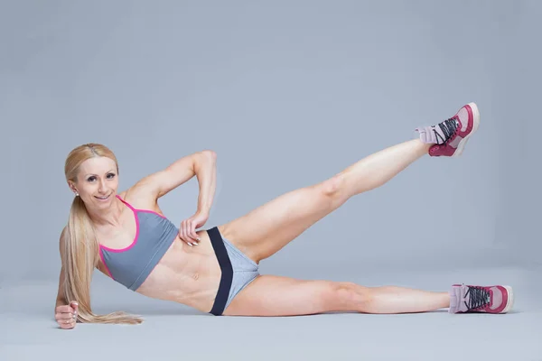 Фітнес дівчина лежить на її боці, піднімаючи ногу, тренує м'язи ніг ізольовані на сірому фоні — стокове фото