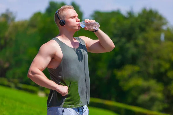 Svalnatý mladý muž ve sluchátkách je pitná voda a odpočinek po běhu v letním parku — Stock fotografie