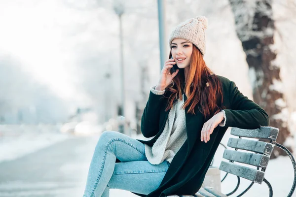 Young woman in winter park talking mobile phone, sms, sitting on the bench — Stock Photo, Image