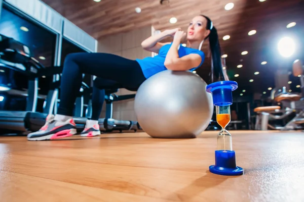 Girl working out at gym with a ball,  in the foreground of an hourglass , time concept to practice — Stock Photo, Image