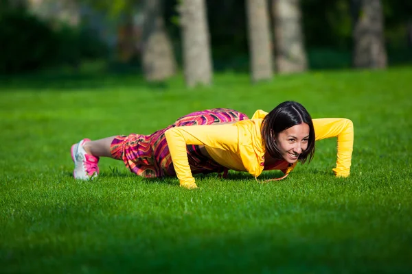 Pers ups uitoefening door jonge vrouw. Meisje uit te werken op gras crossfit sterkte opleiding in de gloed van de ochtendzon — Stockfoto