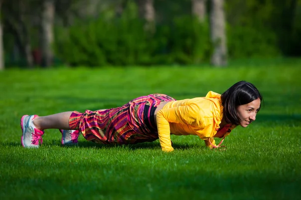 Pers ups uitoefening door jonge vrouw. Meisje uit te werken op gras crossfit sterkte opleiding in de gloed van de ochtendzon — Stockfoto