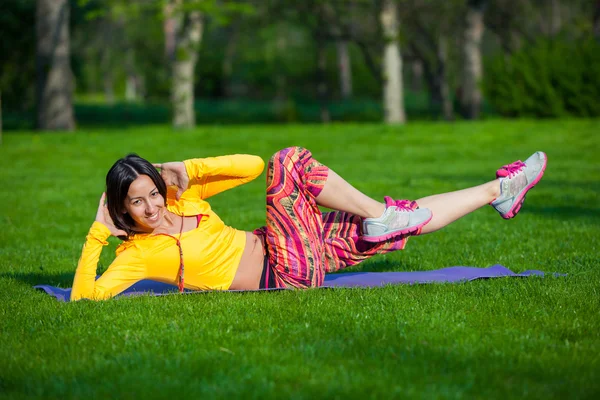 Ejercicio de fitness mujer se sienta fuera durante el entrenamiento de ejercicio crossfit. Chica en forma feliz haciendo crujidos laterales con piernas elevadas mientras sonríe feliz . — Foto de Stock