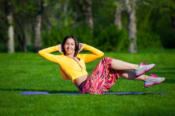 Осуществление фитнес-женщина сидит снаружи во время кроссфит тренировки упражнения. Счастливая подтянутая девушка делает боковые хрусты с высоко поднятыми ногами и счастливо улыбается . — стоковое фото