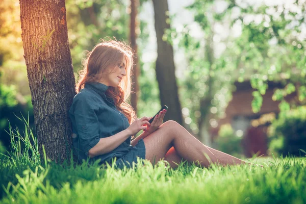 Αρκετά νεαρή καυκάσιος γυναίκα που κάθεται έξω από κάτω από ένα δέντρο που μιλάει στο συσκευή smartphone. Φιλτραρισμένα αποτελέσματα — Φωτογραφία Αρχείου