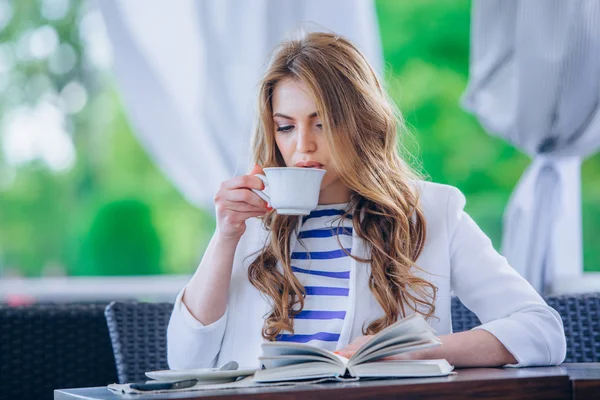 Hermosa joven en la cafetería al aire libre leyendo un libro y beber café. teléfono. Estudiante. mujer de negocios — Foto de Stock