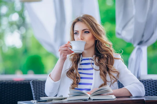 Hermosa joven en la cafetería al aire libre leyendo un libro y beber café. teléfono. Estudiante. mujer de negocios — Foto de Stock