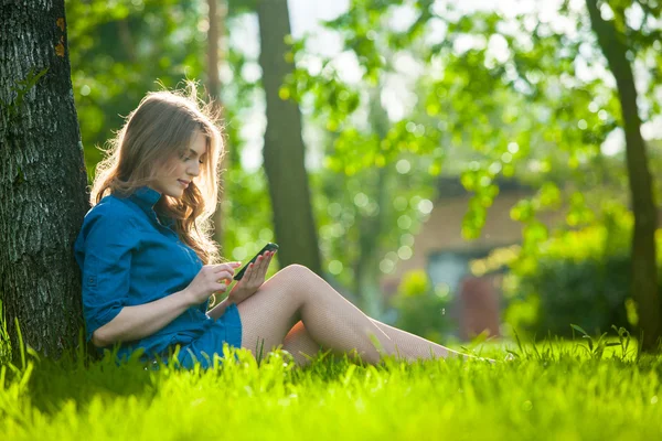 Αρκετά νεαρή καυκάσιος γυναίκα που κάθεται έξω από κάτω από ένα δέντρο που μιλάει στο συσκευή smartphone. Φιλτραρισμένα αποτελέσματα — Φωτογραφία Αρχείου
