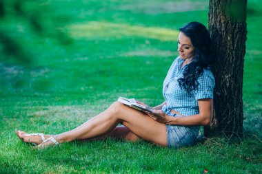 lifestyle, summer vacation, education, literature and people concept - smiling young girl reading book sitting on grass in park