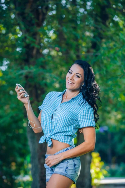 Young hipster stylish beautiful girl listening to music, mobile phone,headphones, enjoying, denim outfit, smiling, happy, cool accessories, having fun, laughing, park — Φωτογραφία Αρχείου