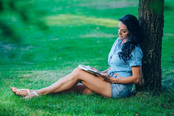 Estilo de vida, férias de verão, educação, literatura e conceito de pessoas - sorridente jovem leitura livro sentado na grama no parque — Fotografia de Stock