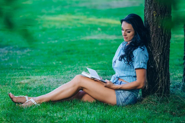 Estilo de vida, férias de verão, educação, literatura e conceito de pessoas - sorridente jovem leitura livro sentado na grama no parque — Fotografia de Stock