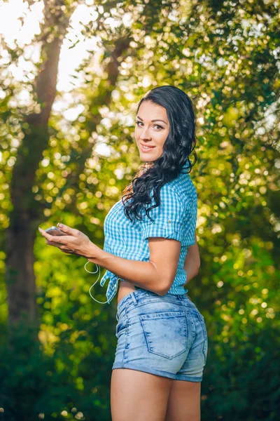 Young hipster stylish beautiful girl listening to music, mobile phone,headphones, enjoying, denim outfit, smiling, happy, cool accessories, having fun, laughing, park — Stock Fotó