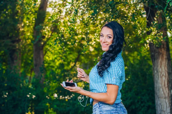 Young hipster stylish beautiful girl listening to music, mobile phone,headphones, enjoying, denim outfit, smiling, happy, cool accessories, having fun, laughing, park — 图库照片