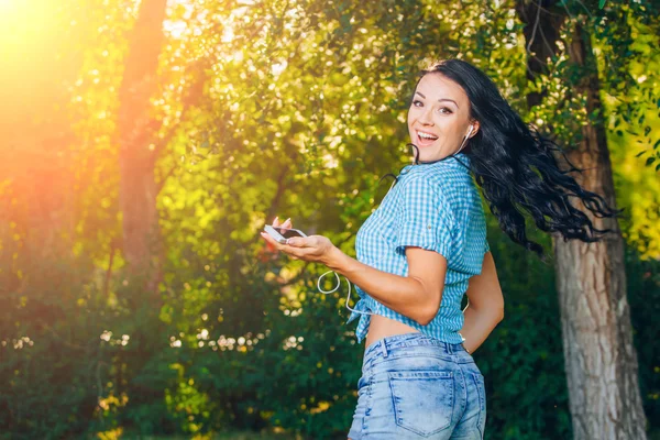 Young hipster stylish beautiful girl listening to music, mobile phone,headphones, enjoying, denim outfit, smiling, happy, cool accessories, having fun, laughing, park — Stock Fotó