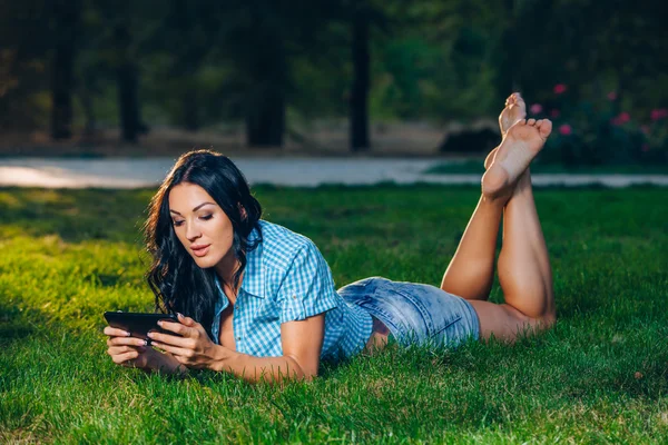 使用 tablet 室外躺在草地上的年轻女子微笑. — 图库照片