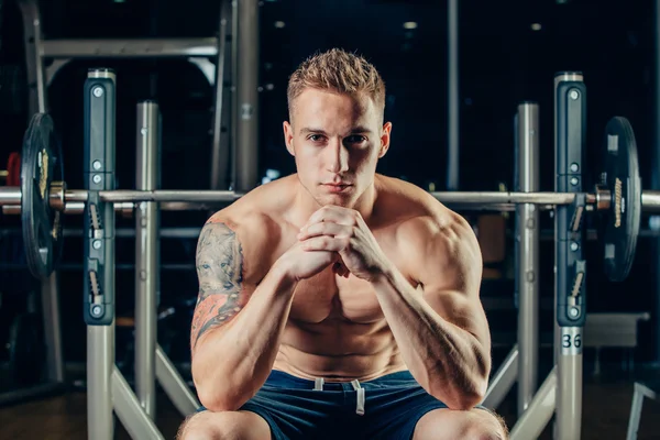 Retrato de cerca de un entrenamiento de hombre musculoso con barra de pesas en el gimnasio. Él está sentado en la vista de marco, pensando —  Fotos de Stock