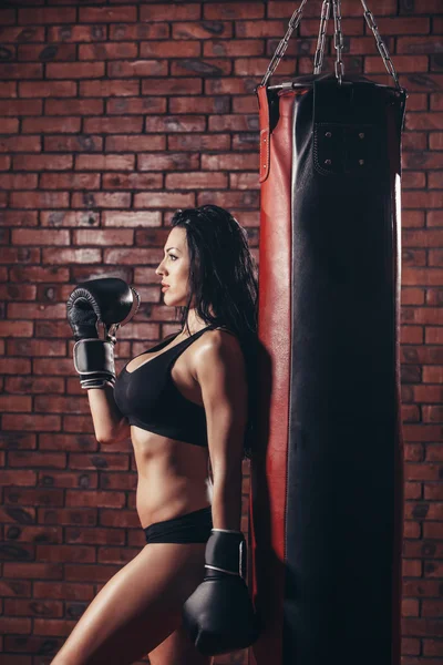 Σέξι κοπέλα με γάντια του μποξ τσάντα punching, στον τοίχο φόντο κόκκινο τούβλο. — Φωτογραφία Αρχείου