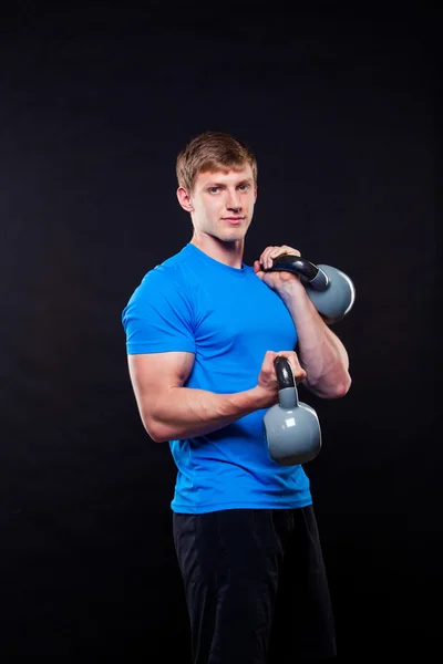 Mladý atletický muž stál s kettlebells na černém pozadí. — Stock fotografie