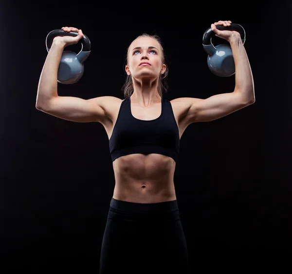 Fitness jovem mulher de pé com kettlebells no fundo preto — Fotografia de Stock