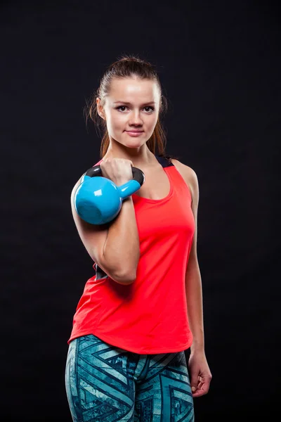 Fitness jovem mulher em camisa vermelha de pé com kettlebells no fundo preto — Fotografia de Stock