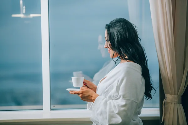 Pierwsze ciepłe ze świeżo parzoną kawą. Piękna młoda kobieta w biały szlafrok, picia kawy i patrząc przez okno — Zdjęcie stockowe